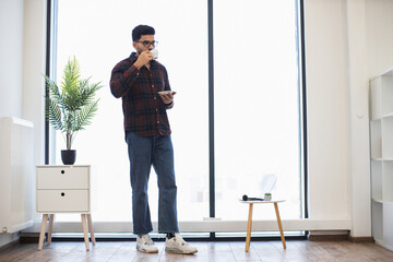 Indian man wearing glasses in casual clothes stands in bright room, enjoys coffee while using...