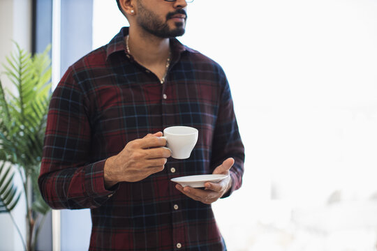 Indian man in casual plaid shirt holding coffee cup with saucer indoors. Relaxed moment enjoying hot beverage. Mid adult male creating calm, warm atmosphere. - Powered by Adobe