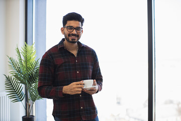 Young Indian man in casual attire smiling while holding coffee in bright office setting. Indoor...