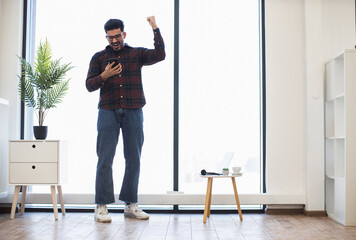 Indian male in casual clothing celebrating success while looking at smartphone in bright modern room. Expresses joy and achievement.