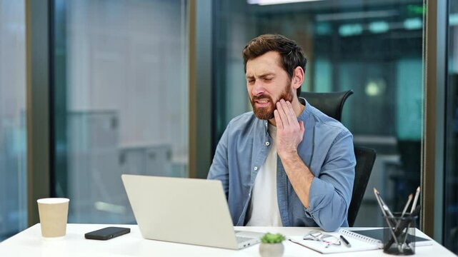 Bearded businessman suffering from toothache in modern office. Unhappy male has a tooth pain, touching cheek. Sick worker with strong tooth pain sitting at workplace.