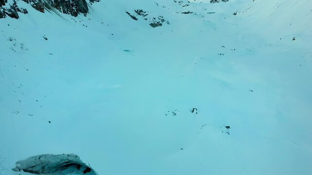 Aerial view of a remote snow covered mountain valley of the Swiss Alps with a pan up motion from a giant glacier cave in Valais, Switzerland to sunlit mountain peaks a cold winter day