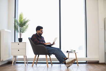 Young Indian man sitting on chair using laptop in bright modern living room. Relaxed work from home...