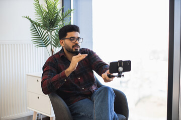 Indian male vlogger recording content at home using smartphone. Casual setting with natural light...