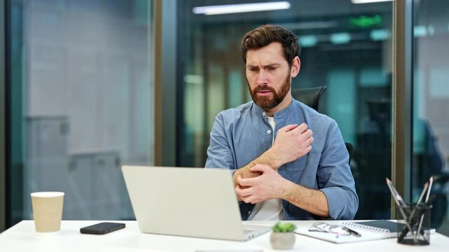 Frustrated businessman experiencing skin irritation and neck pain while working on laptop in modern office. Office worker scratches an itchy area on arm, neck. Businessman has allergies.