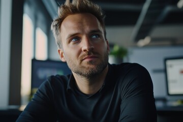 Pensive young Caucasian man in a black shirt, reflecting in an modern office environment with soft lighting.