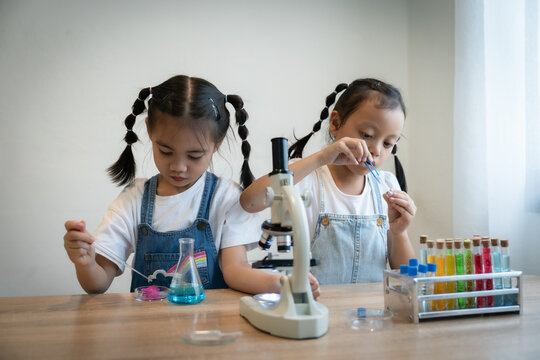 Two young girls are sitting at a table with a microscope and various science experiments. They are both wearing white shirts and denim overalls. The girls are focused on their work