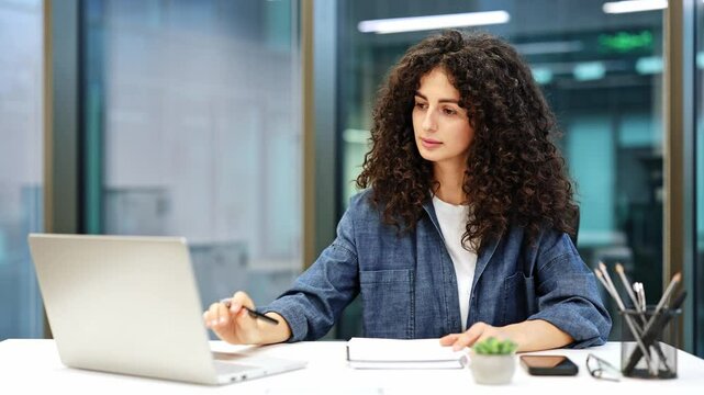 Focused young businesswoman with curly hair works on a laptop and takes notes in modern office. Busy female entrepreneur checks email and writes important information.
