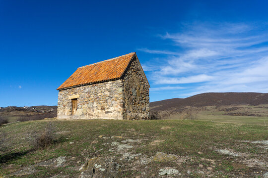 Small shurch on the rock. Wooden door. Orange tiled roof, stone walls. Green grass and bushes. Bright blue sly with clouds. Georgia