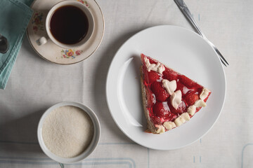 A slice of strawberry cake with cream and strawberry jelly, accompanied by a cup of tea and a sugar bowl on a white tablecloth.