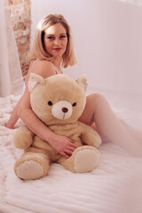 Woman sits on a white bed with a large teddy bear in a softly lit cozy room
