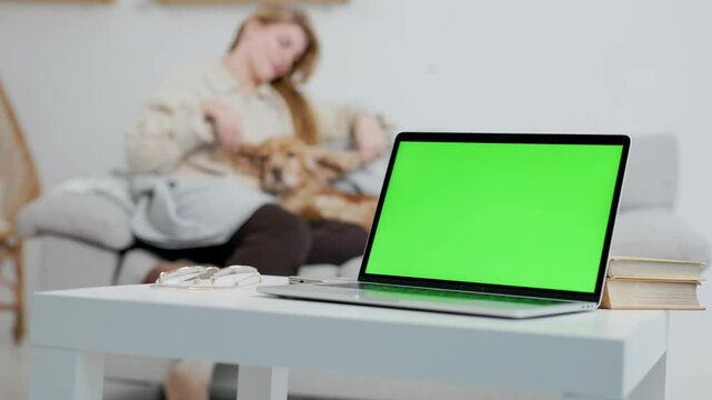 Laptop With Green Mock Up Screen On Table In Living Room. Background- Woman and Her Pet Dog Sitting on Sofa