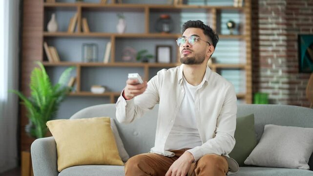 Happy handsome bearded man turns on air conditioner by remote control while sitting on sofa in living room at home. Smiling young adult male presses button of device and turns off cooling in apartment