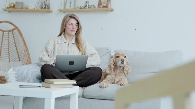 Young Woman in Glasses Using Laptop While Sitting on Couch with Her Dog. Background- Home Interior