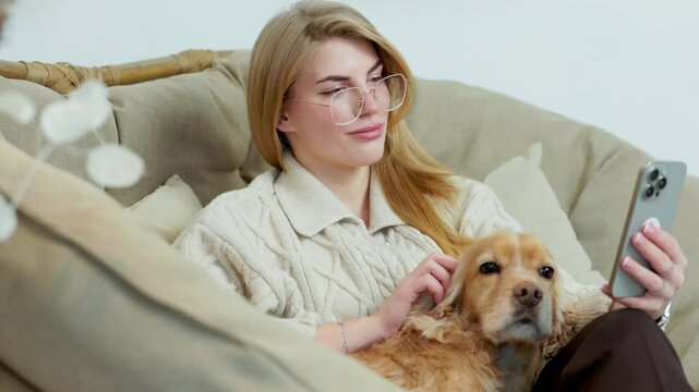 Young Woman Sits in Comfortable Wicker Chair with Her Pet Dog and Flipping Through Her Phone