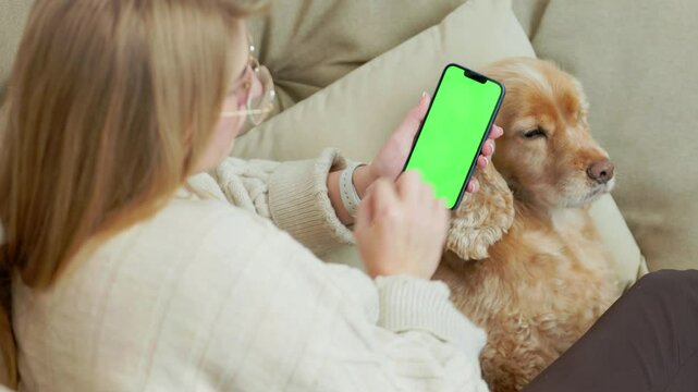 Young Woman Using Green Mock Up Screen Smartphone While Sitting With Her Pet Dog In Cozy Home Room