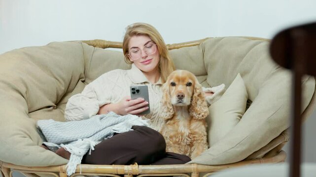 Young Woman Sits in Comfortable Wicker Chair with Her Pet Dog and Scroll Through Phone