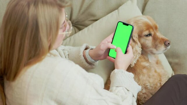 Young Woman Using Green Mock Up Screen Smartphone While Sitting With Her Pet Dog In Cozy Home Room