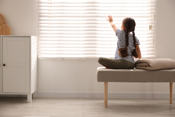 Orphanage concept. Girl sitting on bench and looking through window blinds indoors, back view