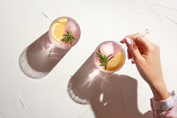 Young woman hand holding glass straw in fresh cold pink lemonade with ice cubes and rosemary....