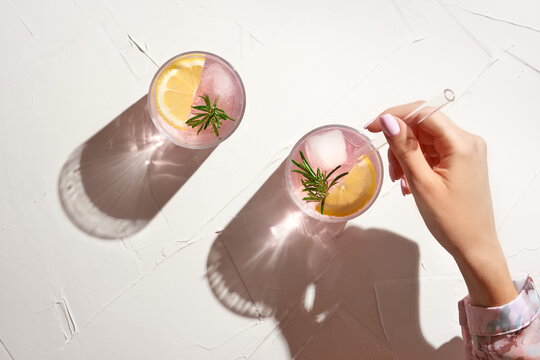 Young woman hand holding glass straw in fresh cold pink lemonade with ice cubes and rosemary. Creative spring, summer concept with bright sunny background with harsh shadows, top view.      