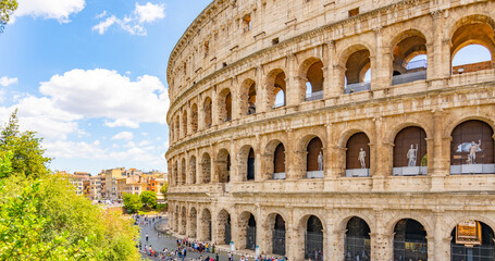 Fototapeta premium The majestic Colosseum stands prominently under a bright sky in Rome. Visitors admire its architecture, while trees and bustling city life create a vibrant atmosphere.