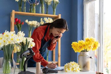 Florist flower shop owner using phone to take orders for her store. She noting order number during smartphone conversation