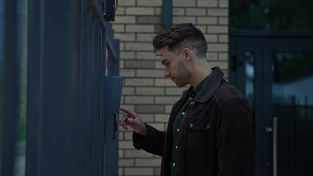 Young man ringing intercom system standing on city street, waiting patiently for access to residential building. Concept of modern technology connecting visitors to secure entrances, slow motion.
