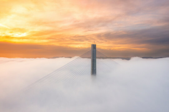 Aerial view of the Queensferry Crossing tower rising through sea fog at sunset in Scotland.