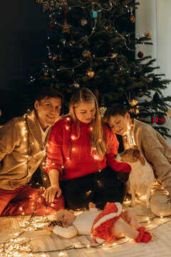 Smiling parents with children and dog wrapped in string light near Christmas tree at home