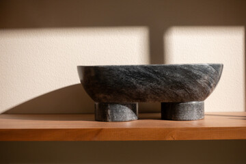 Dark gray granite stone ornament on a wooden shelf against beige wall with shadows from window cast...