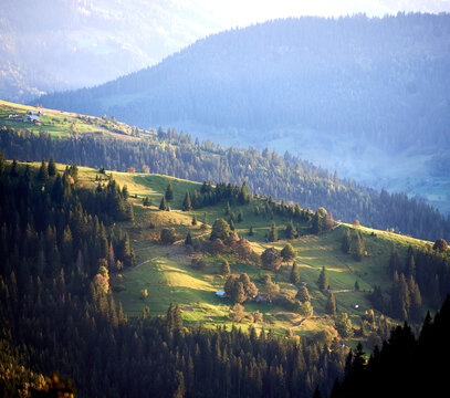 Enchanting view of rolling hills bathed in soft glow of late afternoon light. Patches of sunlight and shadow create mosaic on landscape. Nestled among hills scattered farmhouses and winding paths. - Powered by Adobe