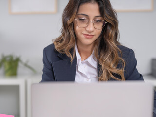 Close up portrait of young happy smiling brunette caucasian business woman sit desk in office using laptop. Stylish female office worker employee looking computer screen. Wear formal jacket, shirt.