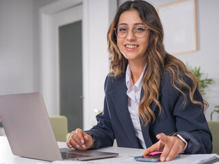 Pretty businesswoman, portrait of young caucasian pretty businesswoman smiling to camera while sitting at her desk at office using laptop holding mobile phone. Wear formal navy blue jacket.