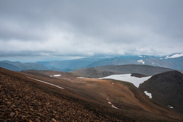 Dark overcast landscape with high pass, wide glacier on stony hill slope and snowy mountain range silhouette in far away in rainy low clouds. Large mountains with snow under gray sky in bad weather.