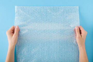 Woman hands holding white transparent plastic air bubble film on light blue table background. Pastel color. Closeup. Material for fragile products packaging. Point of view shot. Top down view.