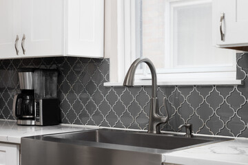 A kitchen faucet detail with a stainless steel apron sink, grey arabesque pattern tile backsplash, and white cabinets and marble countertop.