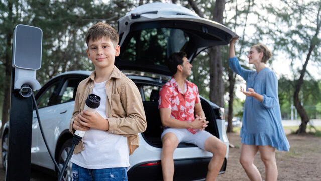 Little boy holding EV charger and point at camera with his family sitting on the trunk in background. Road trip travel with alternative energy charging station for eco-friendly car concept. Perpetual - Powered by Adobe