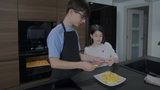 Happy teenage brother wearing apron and sister are tasting freshly baked cookies in a modern kitchen, enjoying the fruits of their labor. Two children eating delicious biscuits baking by them.