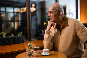  Depressed senior man sitting by the window in the cafe and thinking about something while drinking coffee at evening.