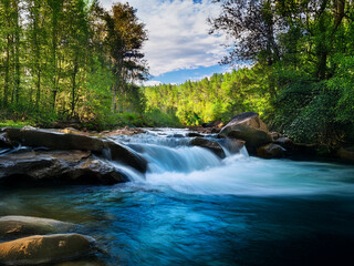 Serene mountain stream cascades over smooth rocks, creating a tranquil scene of vibrant blue water surrounded by lush greenery under a partly cloudy sky.