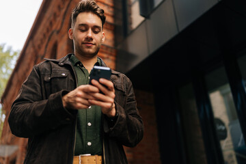 Low-angle view of handsome bearded man standing on city street, using smartphone to check messages,...