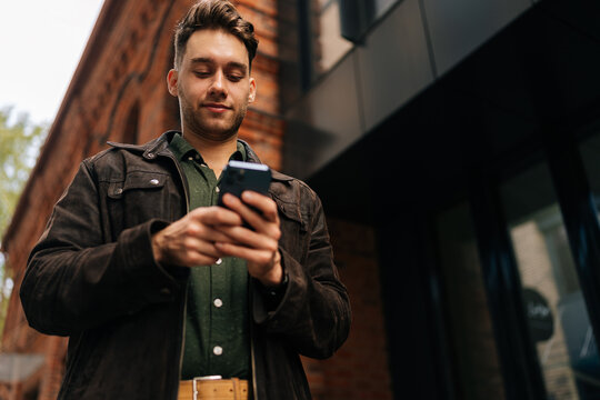 Low-angle view of handsome bearded man standing on city street, using smartphone to check messages, browse internet, stay connected enjoying urban environment. Concept of urban lifestyle. - Powered by Adobe