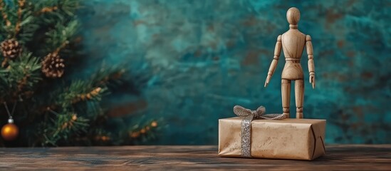 Obraz premium Wooden mannequin stands beside a wrapped gift on a rustic table with a festive Christmas tree in the background.