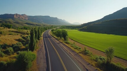 Naklejka premium Scenic aerial view of a winding road through lush green fields and mountains under a clear sky in a serene landscape.