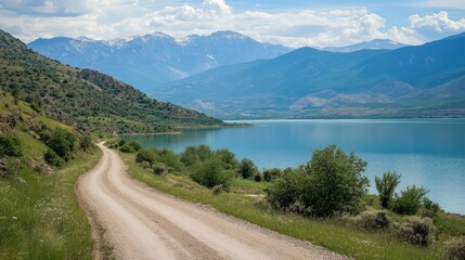 Winding dirt road along serene lake with majestic mountains under blue sky in a picturesque countryside landscape