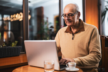 Senior man with eyeglasses using laptop and drinking coffee while sitting in the city cafe at evening.