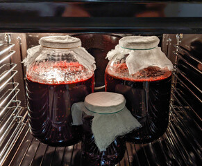 Glass jars filled with homemade wine are ripening inside an oven, covered with breathable cloth to allow fermentation. The rich red liquid glows under the oven light.
