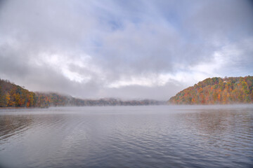 A serene lake scene captures the calm water surrounded by misty fog and trees displaying autumn colors. Gentle ripples spread across the water's surface