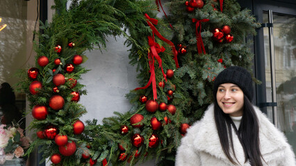 A beautiful, smiling young woman with dark hair, wearing a black hat and a light-colored winter coat, stands near a Christmas wreath. Outdoors. Christmas and New Year's atmosphere.